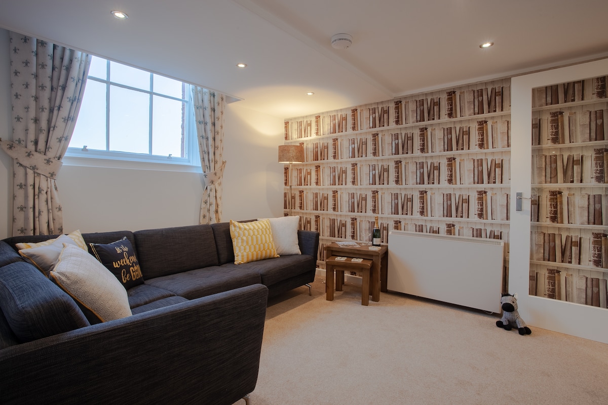 A spacious lounge area features a large, dark grey sectional sofa adorned with textured cushions. Light spills in from a window covered by patterned curtains. A rustic wooden coffee table sits in front of the sofa, complementing the wallpaper with an inviting book pattern.