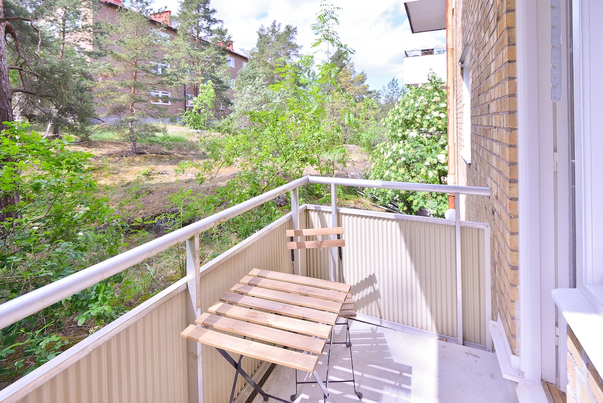 A small balcony features a simple wooden table accompanied by two folding chairs. Lush greenery surrounds the area, providing a sense of privacy and tranquility.