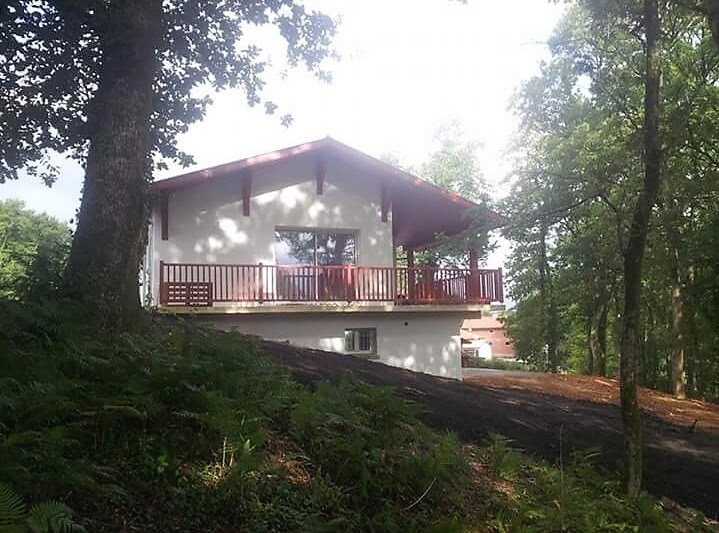 The exterior of a house is surrounded by trees, featuring a covered terrace with wooden railing. The structure includes a spacious window, allowing natural light to illuminate the interior. A sloped landscape with greenery leads to the base of the house.