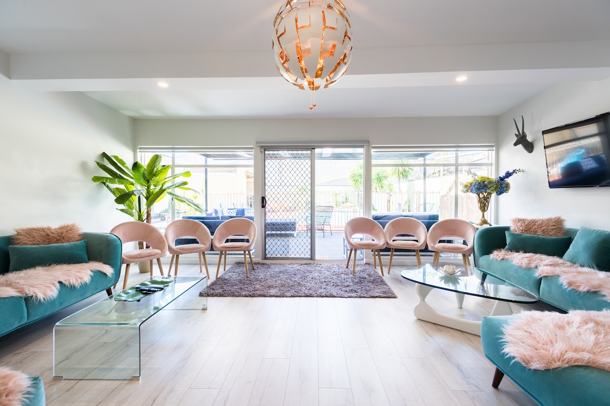 A spacious living area is presented with modern furniture, including plush teal sofas adorned with light pink throws. A stylish glass coffee table sits atop a soft rug, while large sliding doors provide views of the outdoor area, creating a bright and airy atmosphere.