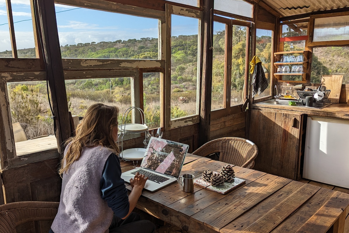 A cozy workspace features a wooden table with a laptop and mug, surrounded by large windows that offer views of the natural landscape. Wooden shelves hold glassware, and a two-tiered serving stand is present. A rustic kitchen area is visible in the background.