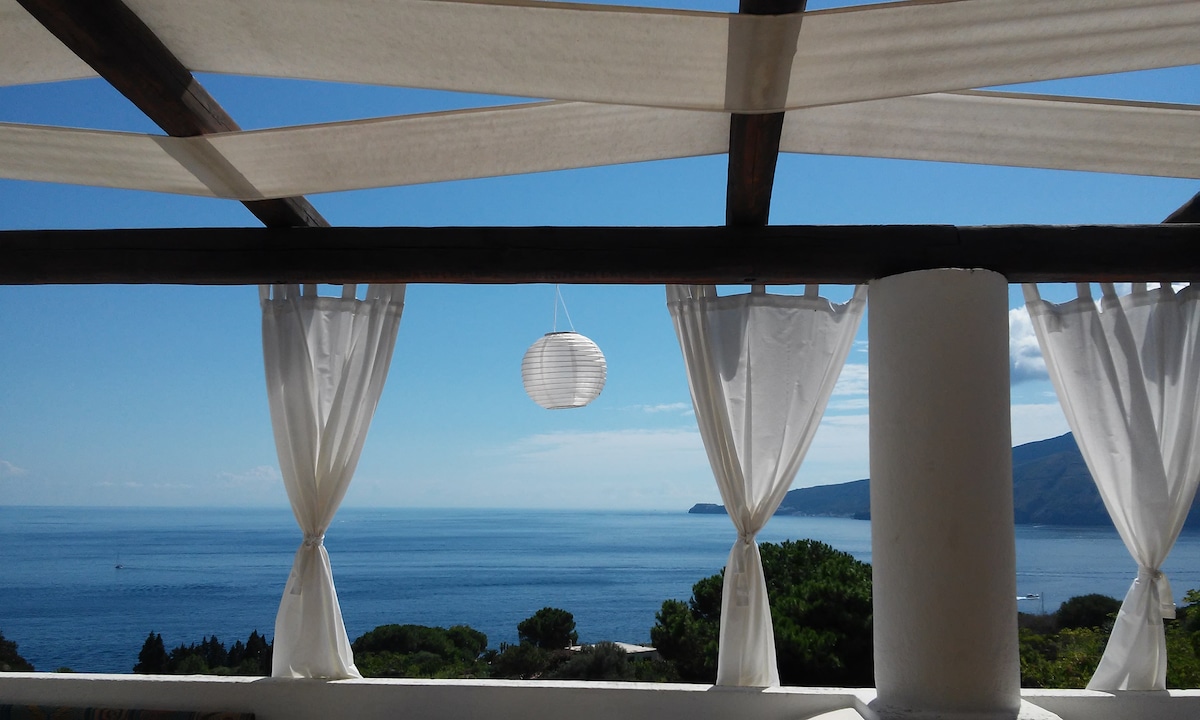 A bright, open terrace offers sweeping views of the serene sea, framed by flowing white curtains. A decorative lantern hangs from the ceiling, complementing the tranquil atmosphere and inviting natural light.
