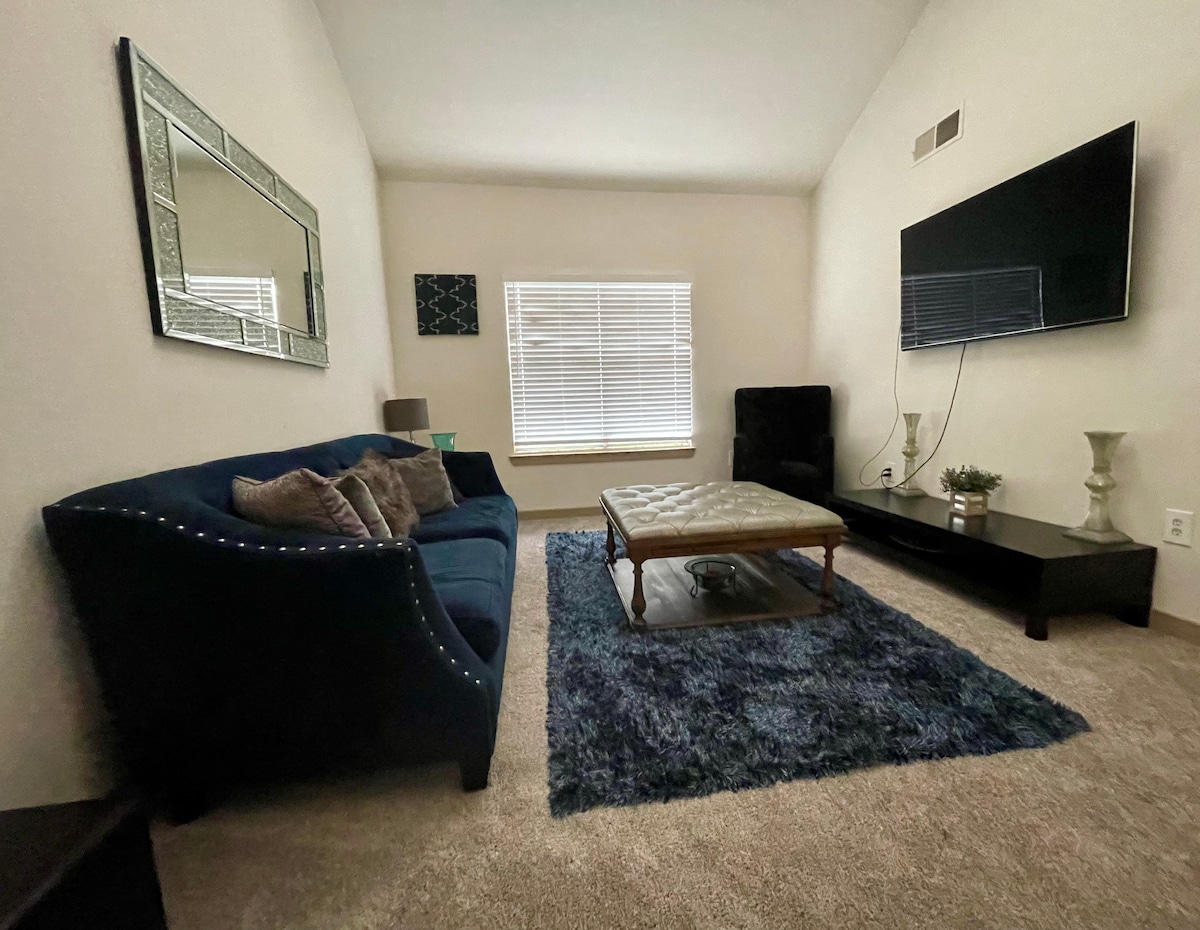 A cozy living area features a plush navy sofa adorned with decorative cushions, positioned beside a stylish coffee table. A large flat-screen television is mounted on the wall, while light streams in through a window dressed with blinds, illuminating the soft carpet.