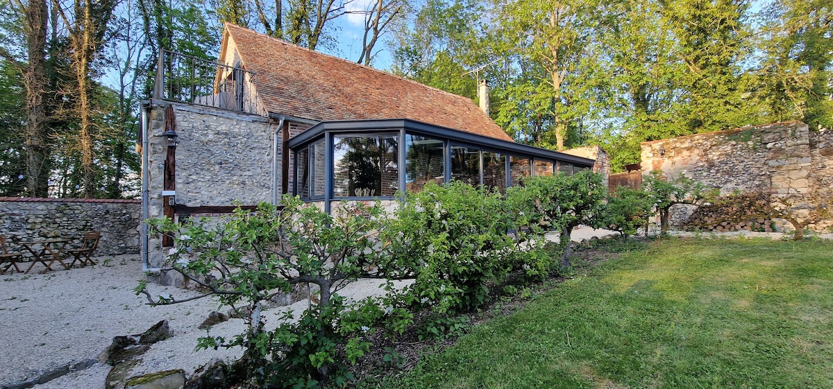The exterior features a stone house with a traditional roof, complemented by a modern glass veranda. A lush garden surrounds the property with neatly trimmed shrubs and grassy areas, providing a serene outdoor space. Trees and greenery create a natural backdrop.