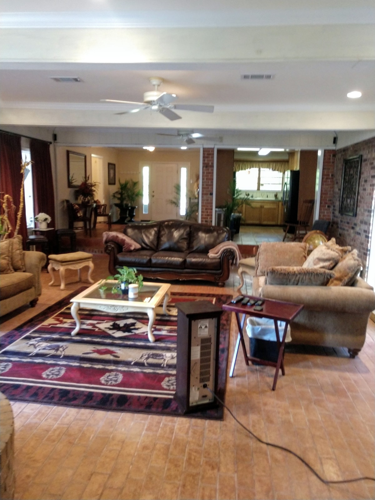 A spacious living area is furnished with a dark leather sofa and two armchairs. A coffee table with a small plant sits centrally, surrounded by a soft area rug. Natural light floods in through large front windows, illuminating the open layout.