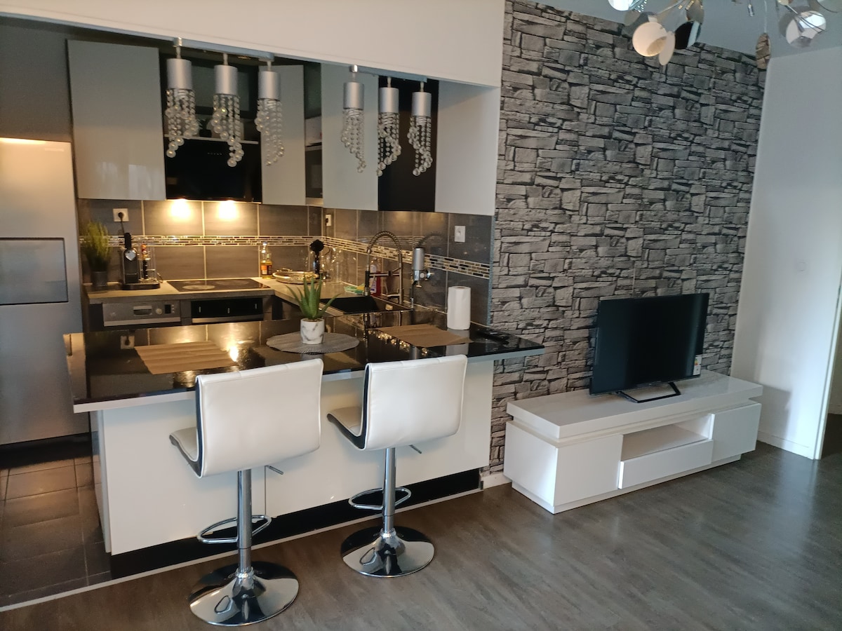 A modern kitchen is showcased with sleek cabinetry and a stone accent wall. High-top bar stools accompany a kitchen island, while stylish pendant lights hang above. A flat-screen TV is positioned on a low, white media console, contributing to the contemporary aesthetic.