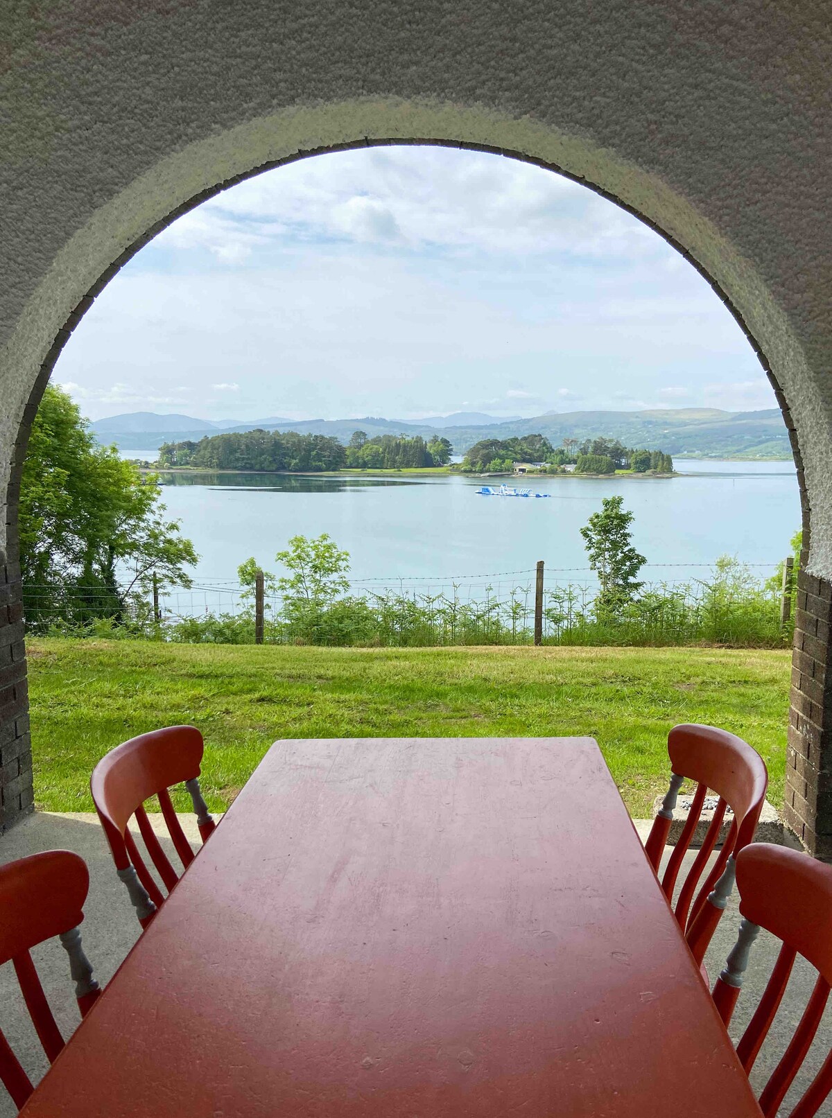 A view from a covered porch features a large red timber table and four chairs, overlooking Kenmare Bay. Lush greenery frames the scene, with distant hills visible across the water and boats dotting the bay.