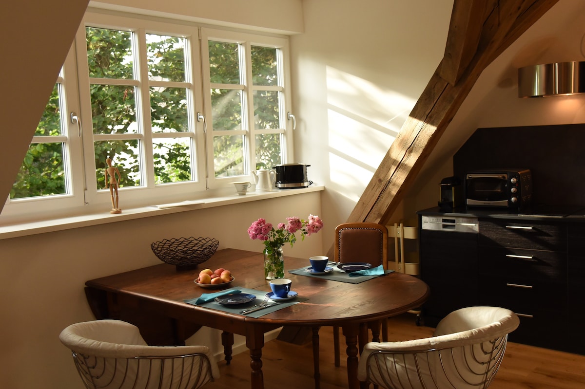 A bright dining area features a wooden table set for two, complemented by woven chairs. A bouquet of flowers sits at the center of the table, while natural light filters through multi-pane windows, enhancing the inviting atmosphere. The kitchenette is visible, equipped with modern appliances.