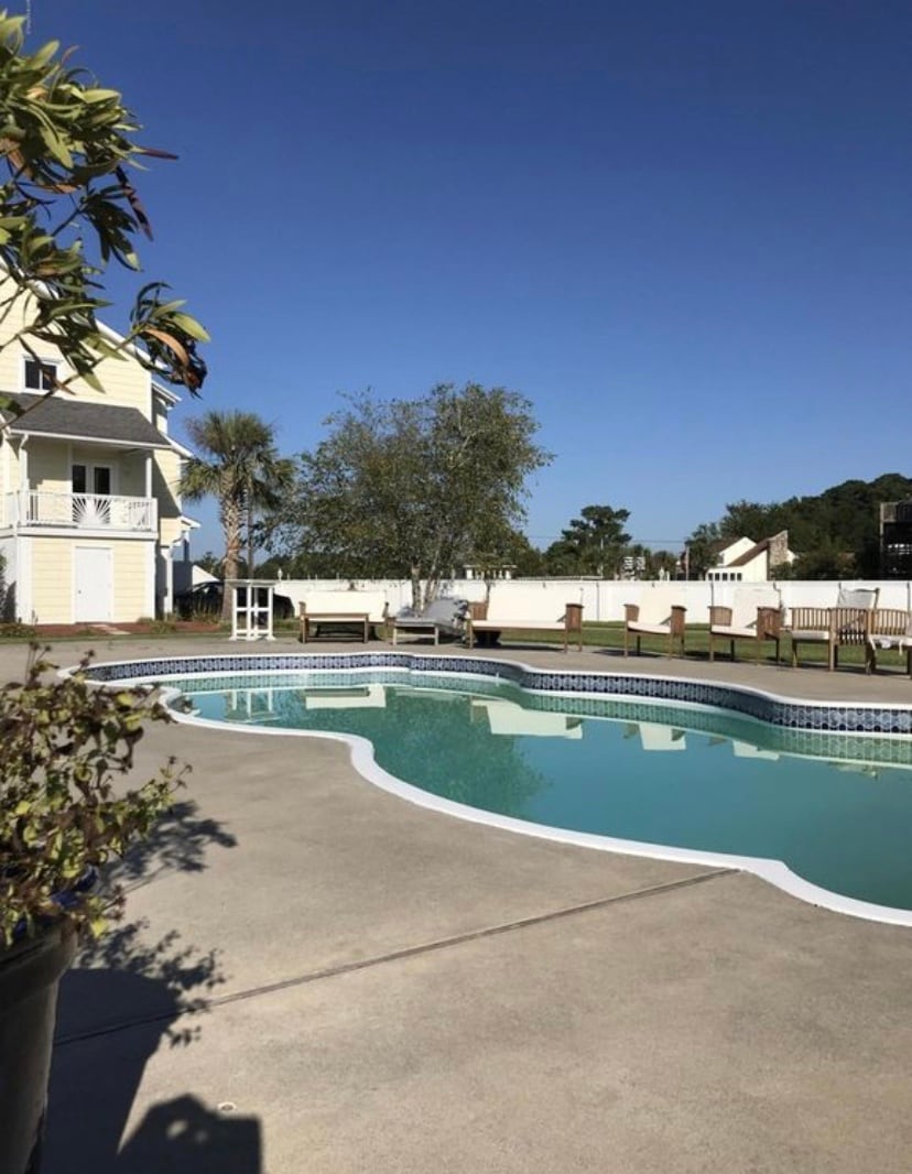 A swimming pool is framed by a spacious patio area, featuring multiple lounge chairs arranged along the edge. The water is clear and reflective under a bright blue sky, with surrounding palm trees and greenery enhancing the outdoor setting.