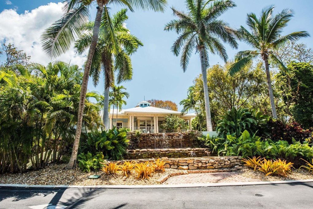 A lush entrance is framed by tall palm trees and vibrant tropical foliage. The pathway leads to a welcoming clubhouse, showcasing a blend of greenery and colorful landscaping against a clear sky.