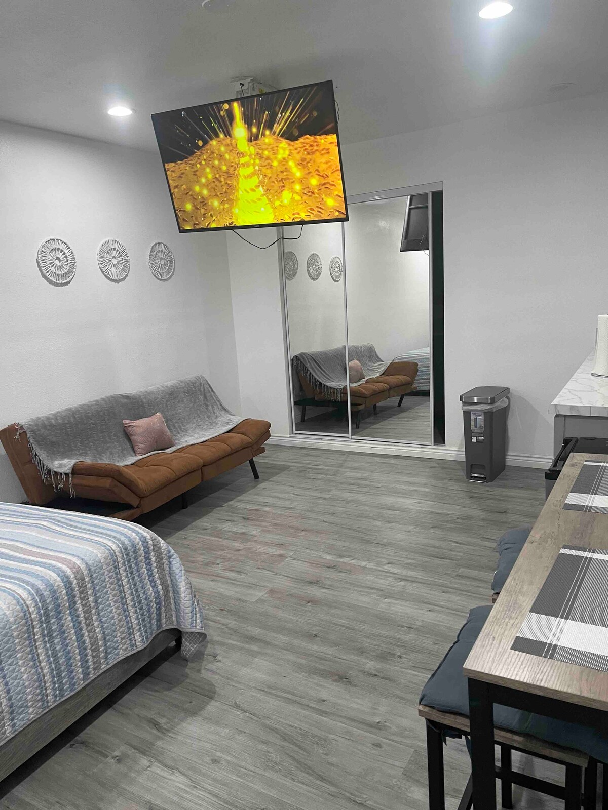 A studio space features a double bed and a sofa bed, complemented by a wall-mounted television. A large mirrored closet is visible. The floor is covered with light-colored wood, and a dining table with chairs occupies one corner, providing a functional area for meals.