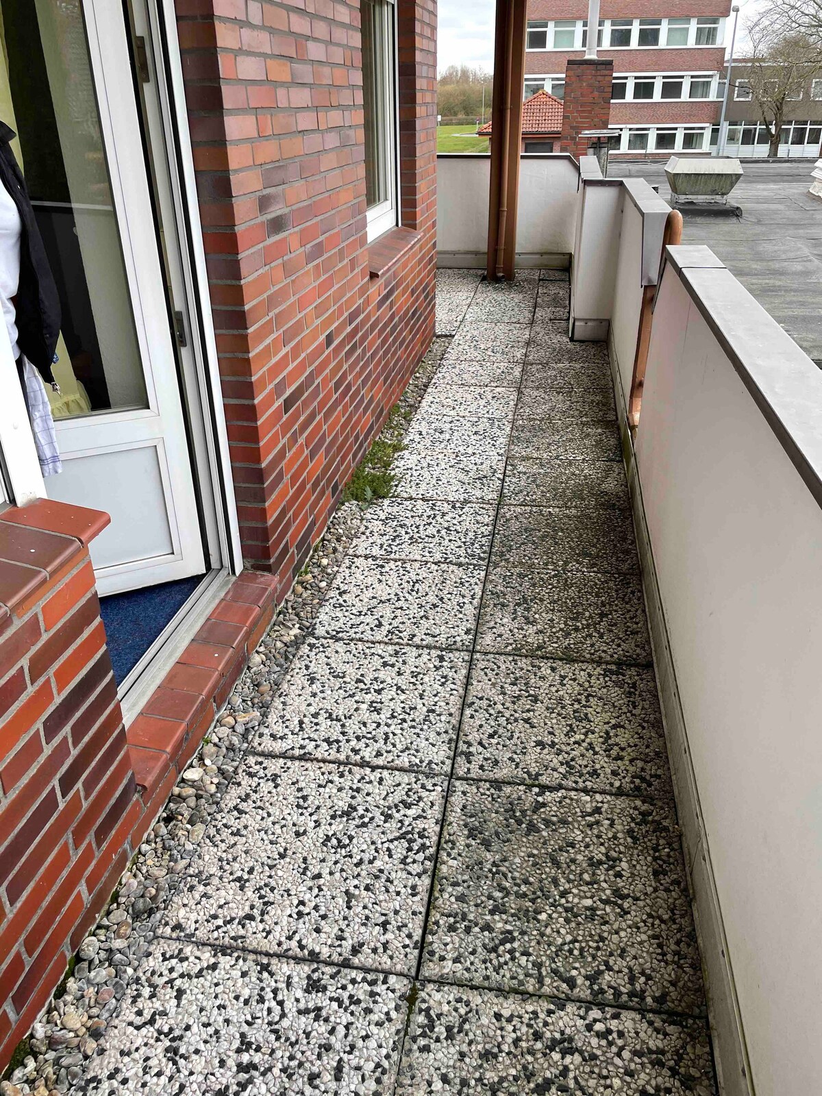 A narrow balcony is displayed, featuring a paved surface with a mix of stone textures and colors. Brick walls line one side, while a glass door leads to the interior of the apartment. A glimpse of greenery and buildings is visible in the background.
