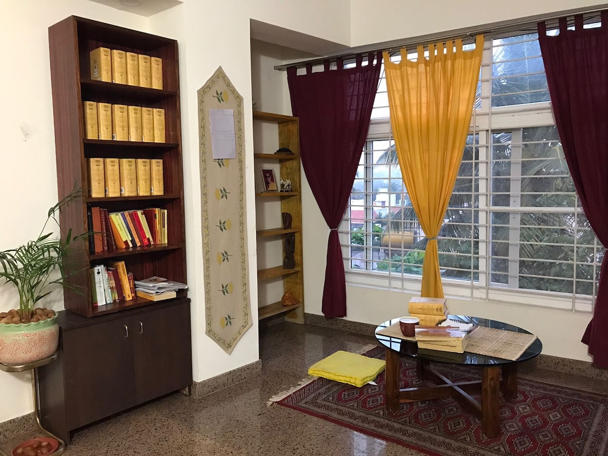 A welcoming corner features a wooden bookshelf filled with books, complemented by a round table on a patterned rug. Bright yellow and maroon curtains frame a window, allowing natural light to illuminate the space, while a potted plant adds a touch of greenery.