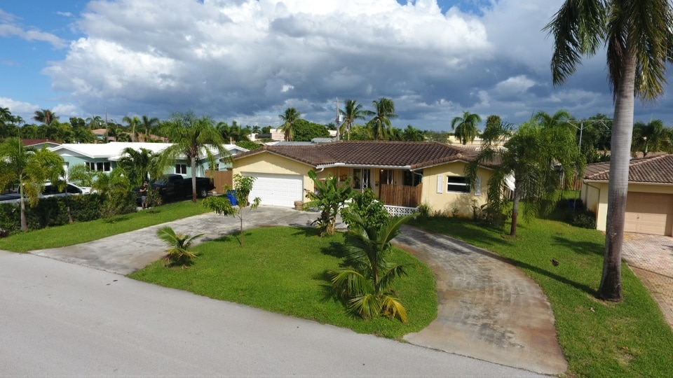 The image showcases a single-story home set on a spacious lawn, surrounded by palm trees. The driveway leads to a garage on the right. A partly cloudy sky forms the backdrop, adding depth to the serene neighborhood setting.