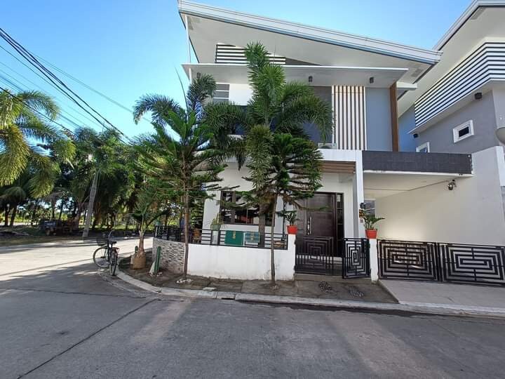 The exterior of a modern villa is presented, showcasing a well-maintained facade with vertical accents. Lush palm trees are arranged symmetrically along the entryway. A pathway leads to the entrance, while a calm street scene is visible nearby.