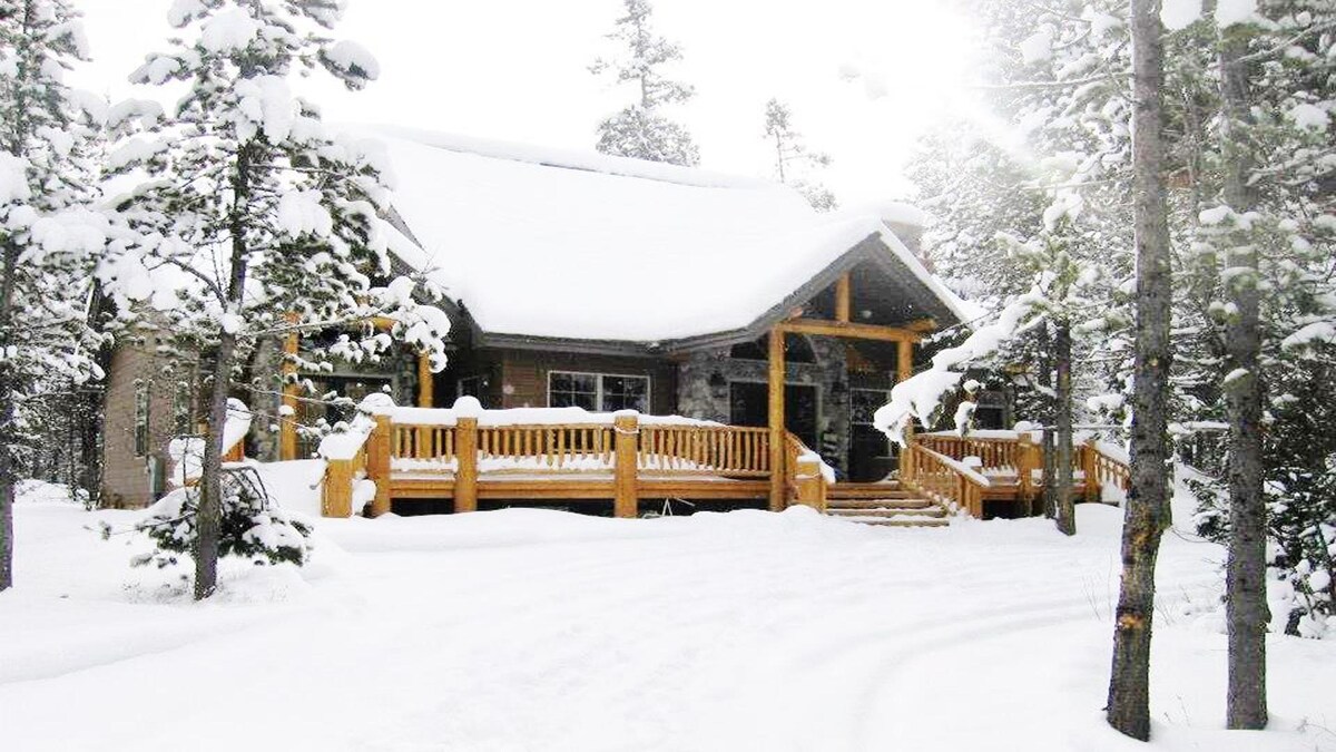 A cabin entrance is framed by snow-covered trees, creating a serene winter landscape. Wooden steps and a deck lead to the front door, showcasing natural wood detailing. The surrounding snow blankets the ground, enhancing the tranquil atmosphere of the forested area.