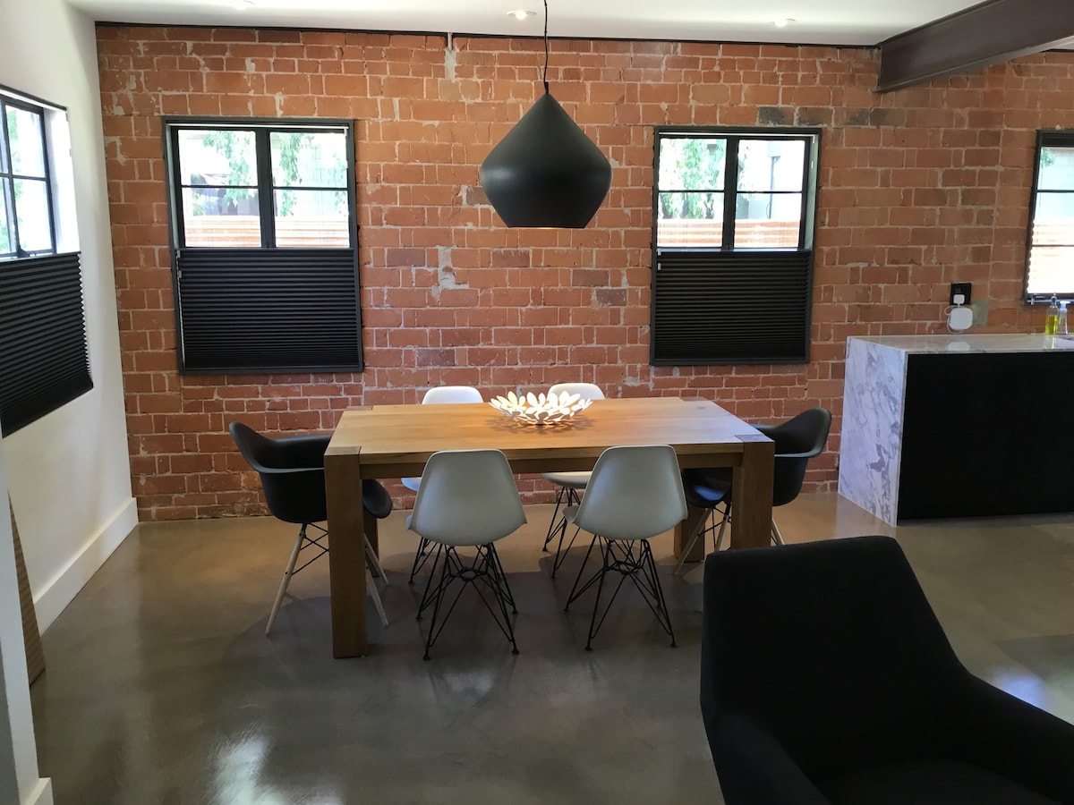 A modern dining area features a large wooden table surrounded by six stylish chairs. The space is adorned with exposed brick walls and large windows, allowing natural light to fill the room. A unique centerpiece sits atop the table, enhancing the contemporary design.