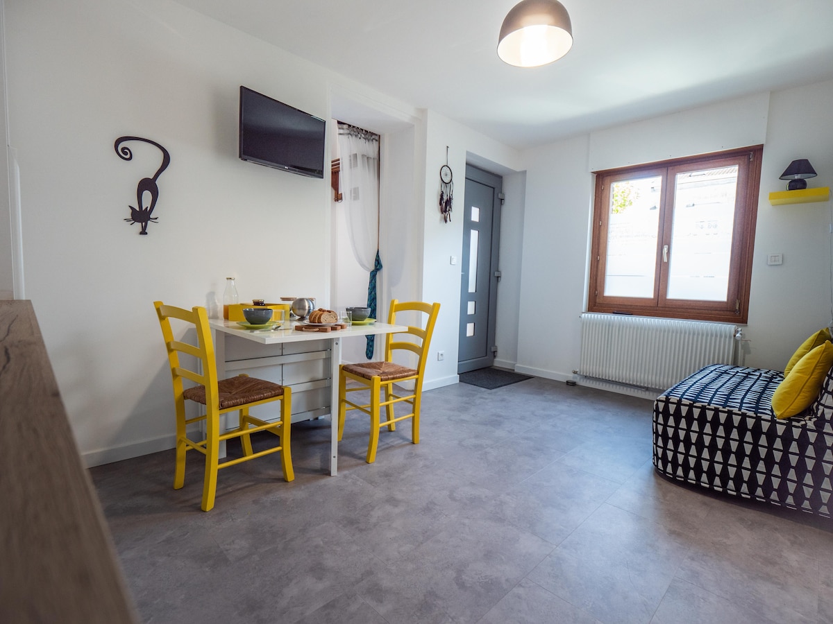 A dining area features a white table paired with four yellow chairs, set with breakfast items. A flat-screen TV is mounted on the wall. Natural light enters through a large window, illuminating the room. A comfortable seating space is arranged with a patterned cushion near the door.