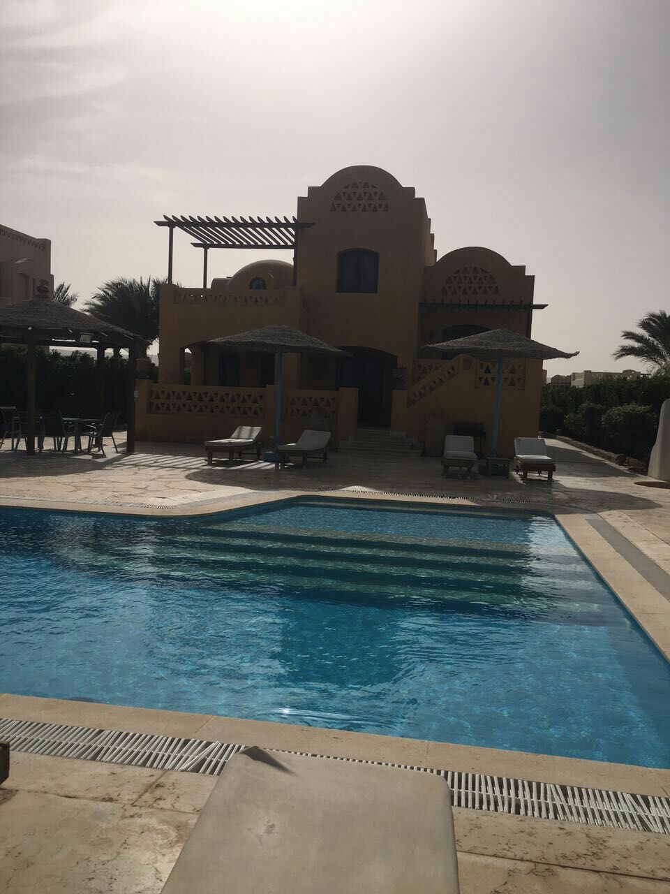 A private pool features crystal-clear water, framed by loungers and umbrellas. The villa's warm-toned facade with intricate architectural details can be seen in the background, surrounded by lush greenery. Multiple levels of the villa are visible, showcasing an inviting outdoor space.