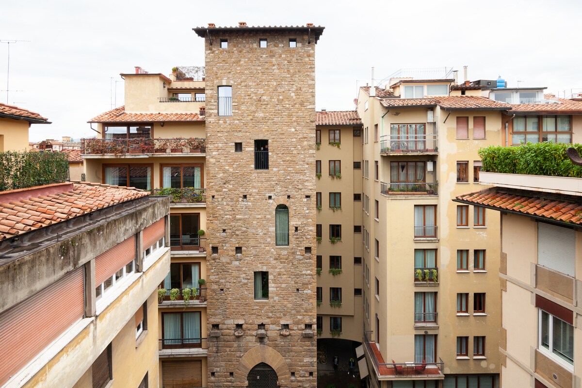 A stone medieval tower stands prominently among modern buildings, showcasing its historic architecture. The roof features terracotta tiles, and several balconies adorned with greenery can be seen on adjacent structures. The sky is overcast, giving a muted backdrop to the scene.