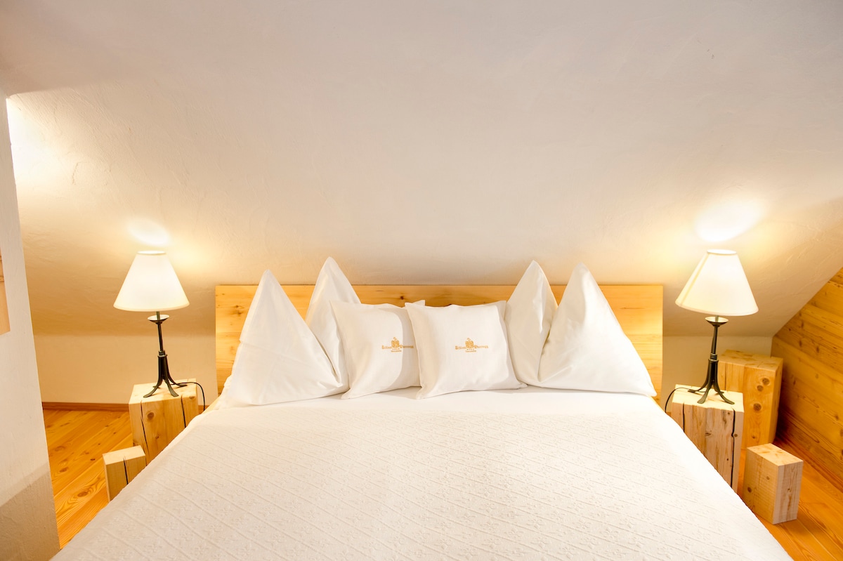 The sleeping area features a spacious bed with white linens and decorative pillows arranged neatly against a wooden headboard. Soft lighting from two bedside lamps creates a warm ambiance, while the angled ceiling adds a cozy character to the room.