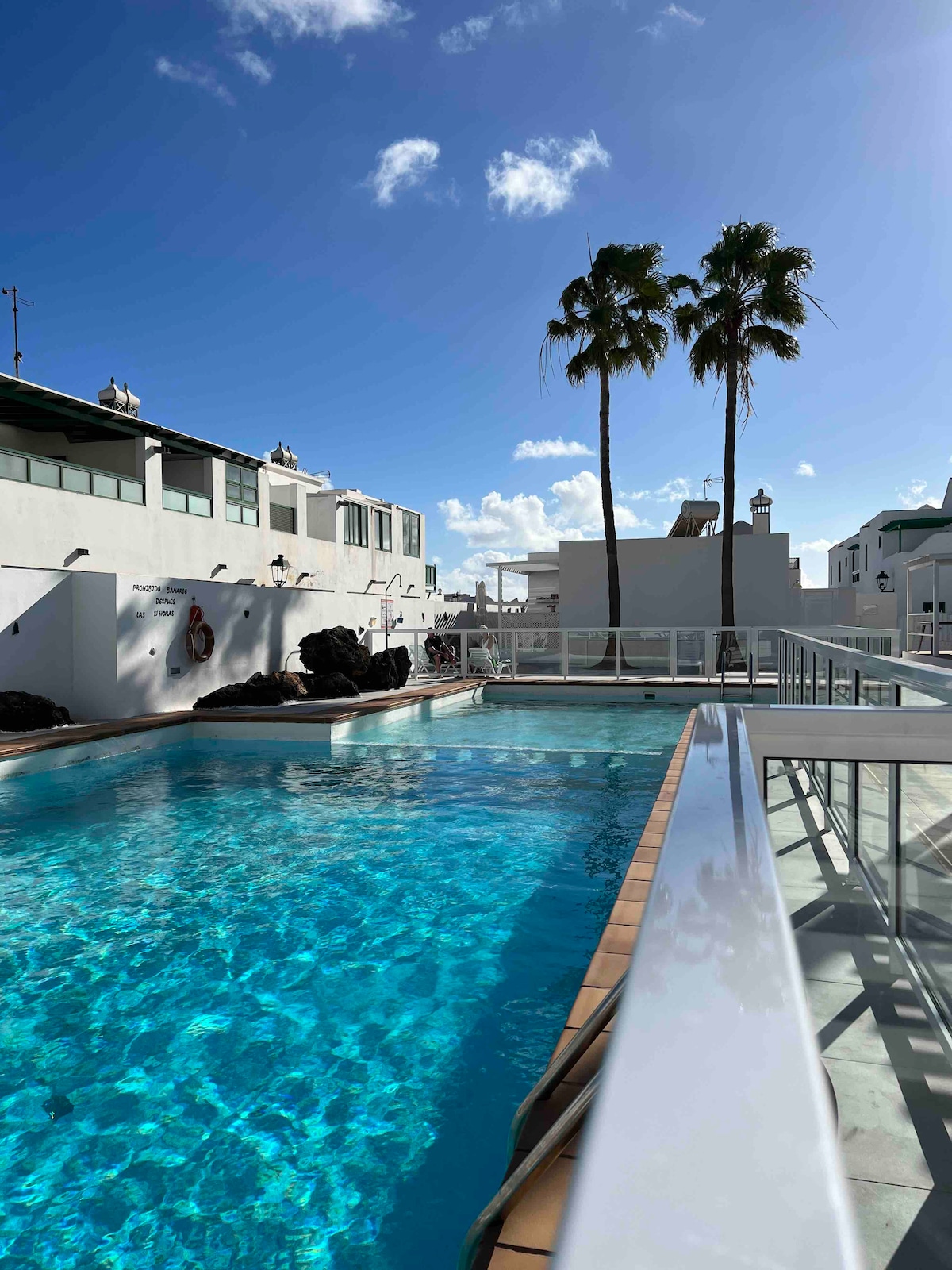 A clear swimming pool glistens under a bright sky, framed by palm trees. Surrounding white buildings are partially visible in the background, reflecting a clean and modern design. The pool area is well-maintained, inviting relaxation and enjoyment in a tranquil setting.