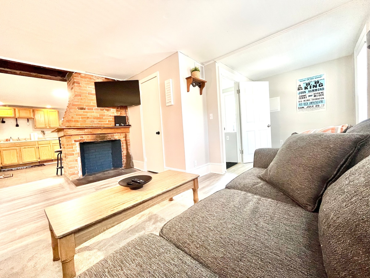 A spacious living area features a comfortable gray couch positioned beside a wooden coffee table. A brick fireplace provides a focal point, while a flat-screen television is mounted on the wall. Natural light enters through nearby windows, enhancing the inviting ambiance.