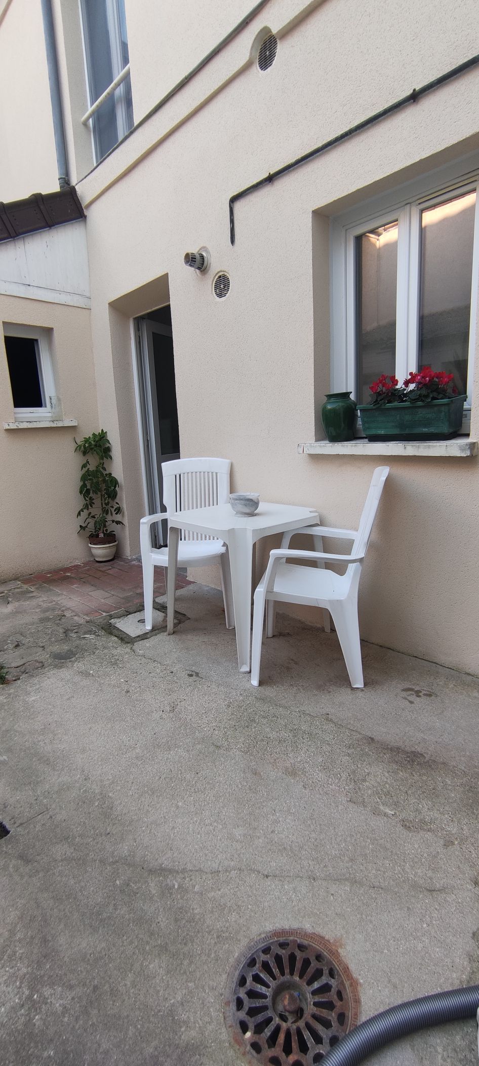 A small outdoor terrace is shown, featuring a white table and two matching chairs placed against a neutral wall. Flower pots with bright red blooms are positioned on the windowsill, enhancing the simple, functional space for relaxation.