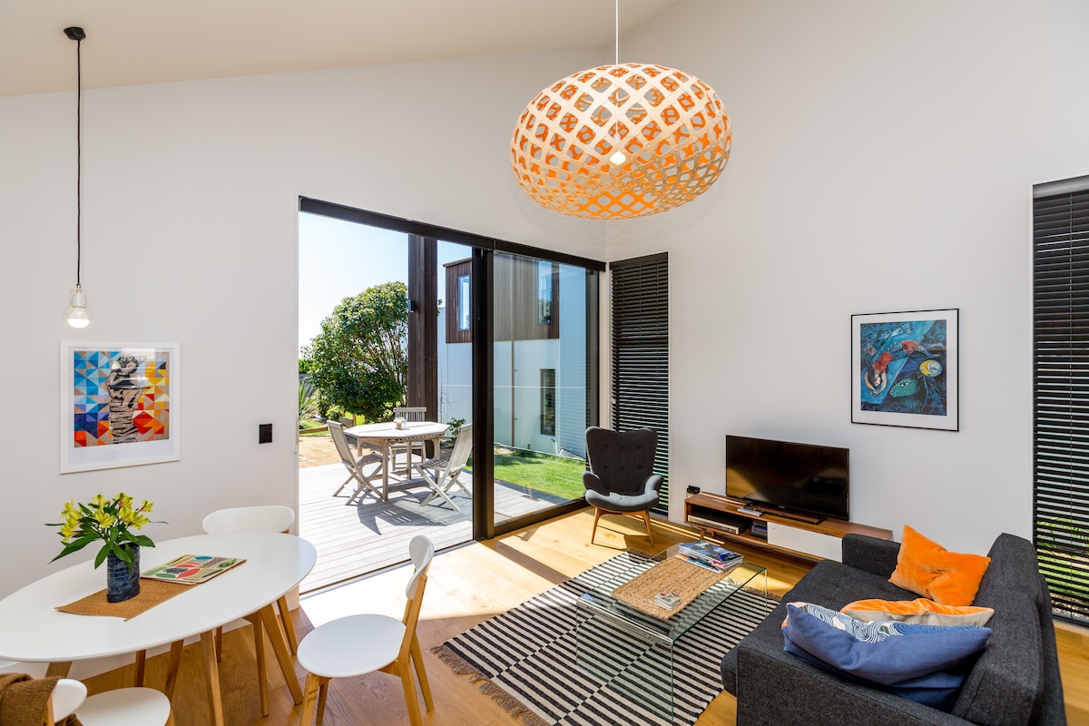The living space features an open layout with large glass doors allowing natural light. A round dining table is paired with white chairs, while a cozy sofa, complemented by a vibrant lamp and artwork, creates a relaxed atmosphere. The outdoor deck is visible beyond.