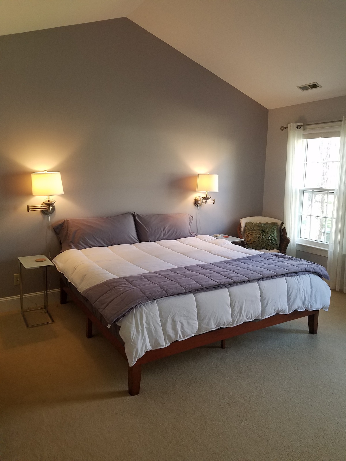A spacious bedroom features a king-size bed with a light purple quilt and white bedding. Two wall-mounted lamps provide soft lighting on either side of the bed. A comfortable chair adds to the inviting space, while natural light filters through the window adorned with light curtains.