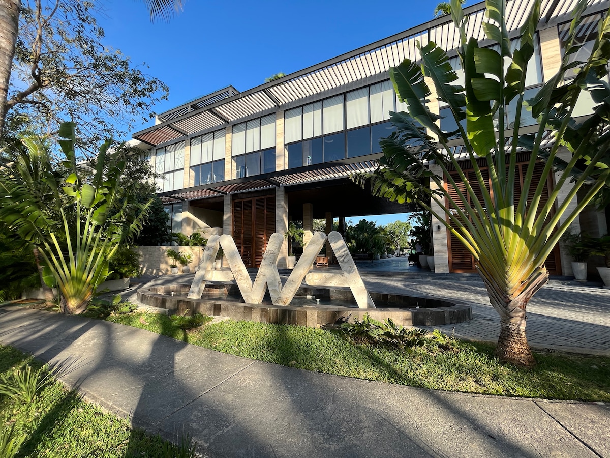 The entrance to a modern building is showcased, featuring large glass windows and a welcoming water feature. Lush greenery surrounds the structure, with tall palm trees and well-maintained landscaping creating an inviting outdoor space.