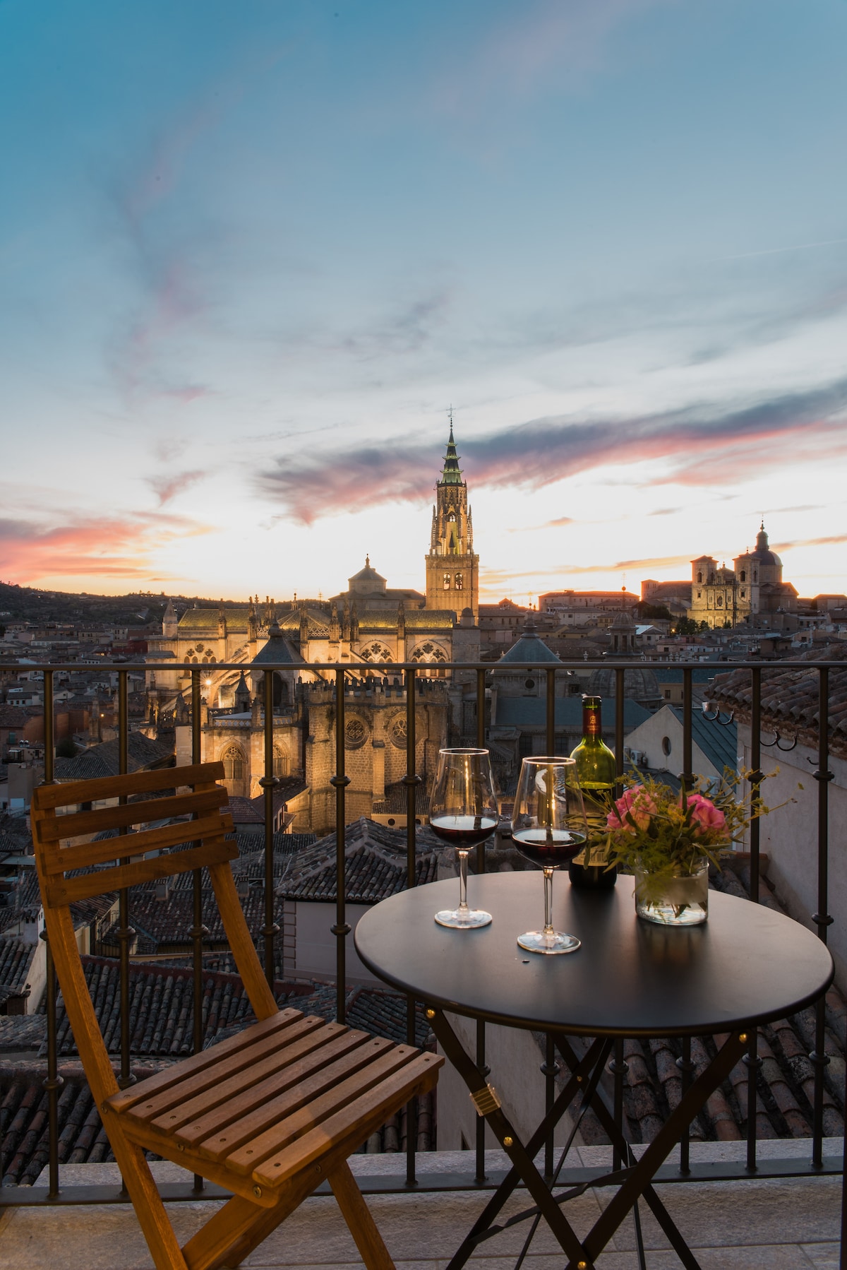 A small balcony features a round table with two glasses of red wine and a decorative vase of flowers. A wooden chair is nearby. The stunning view showcases the city skyline and the lit Cathedral against a colorful sunset sky.