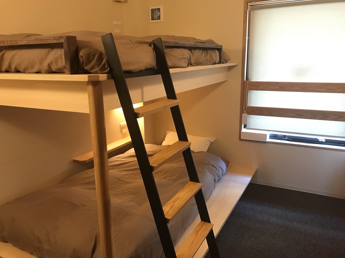 A guest room features a lofted sleeping area above a spacious lower bed, with a wooden ladder providing access. Natural light enters through a window adorned with wooden blinds, casting a soft glow on the warm-colored walls.
