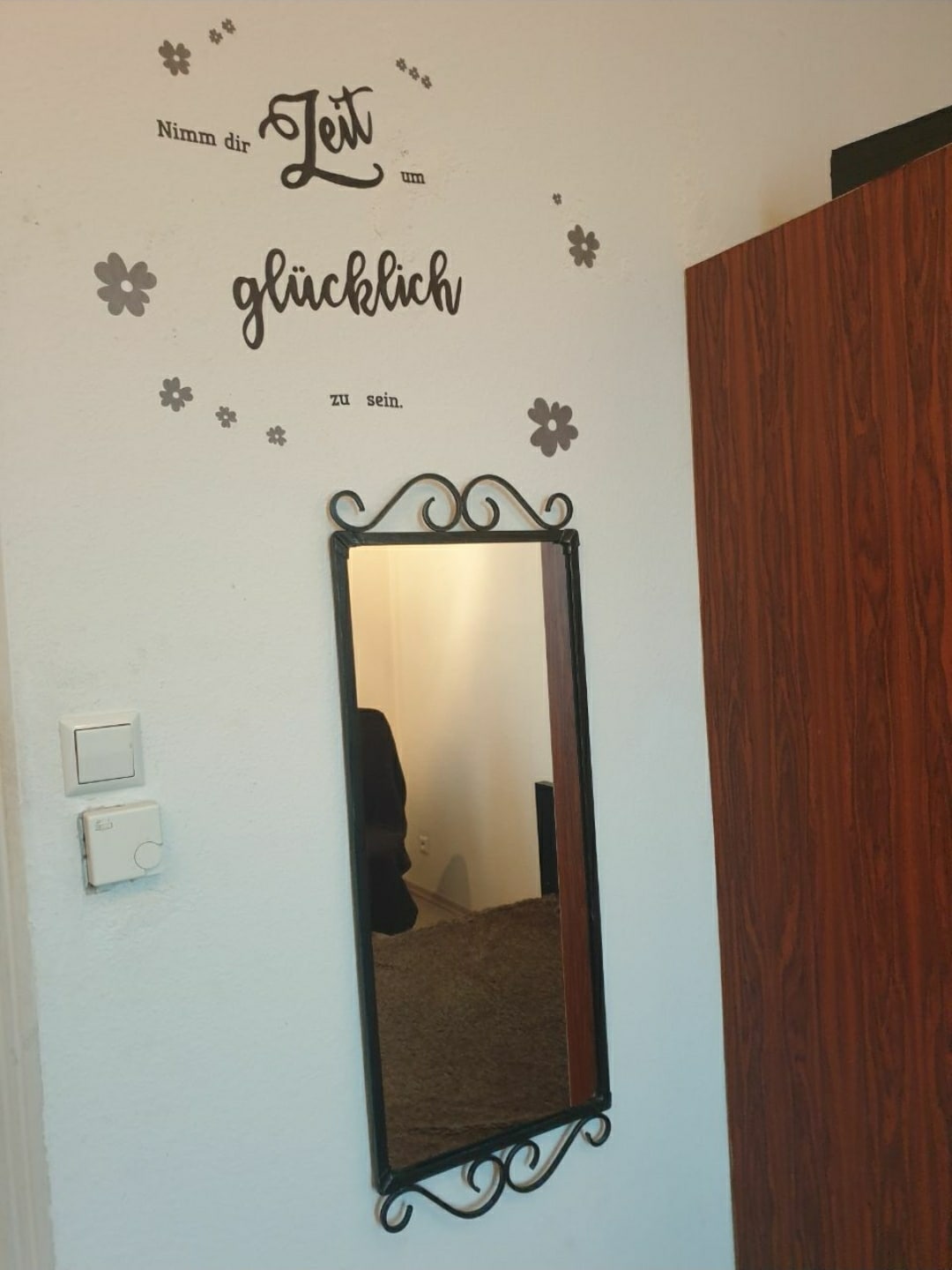 A wall-mounted mirror is framed in black iron, providing a stylish touch to the space. Above the mirror, a decorative quote is displayed on the wall, accompanied by floral designs. A wooden cabinet with a rich brown finish is positioned nearby.