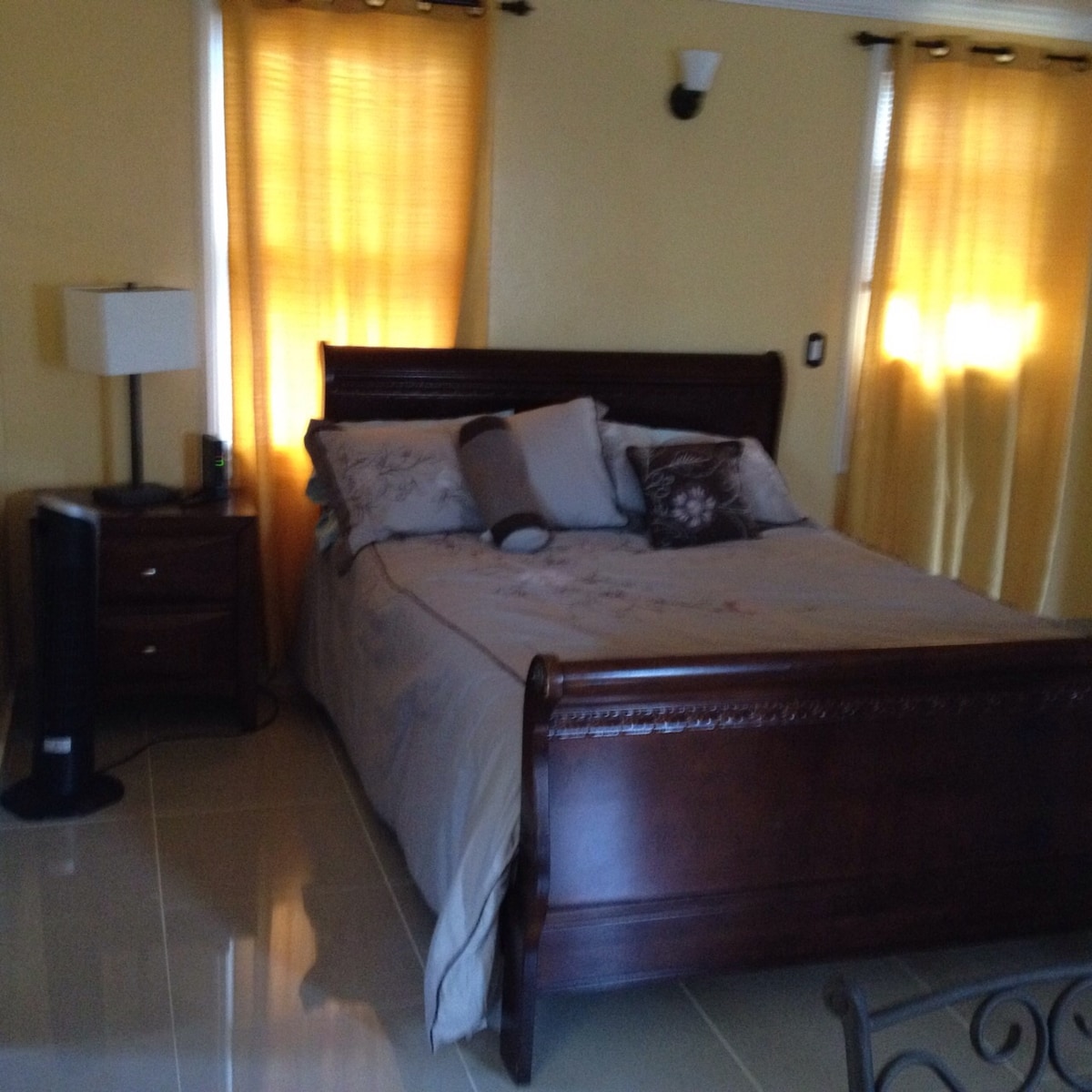 A spacious bed with a light-colored blanket and decorative pillows is positioned against a warm yellow wall. Natural light filters through sheer curtains, creating a soft glow. A bedside table with a lamp is situated next to the bed, complementing the minimalist design.