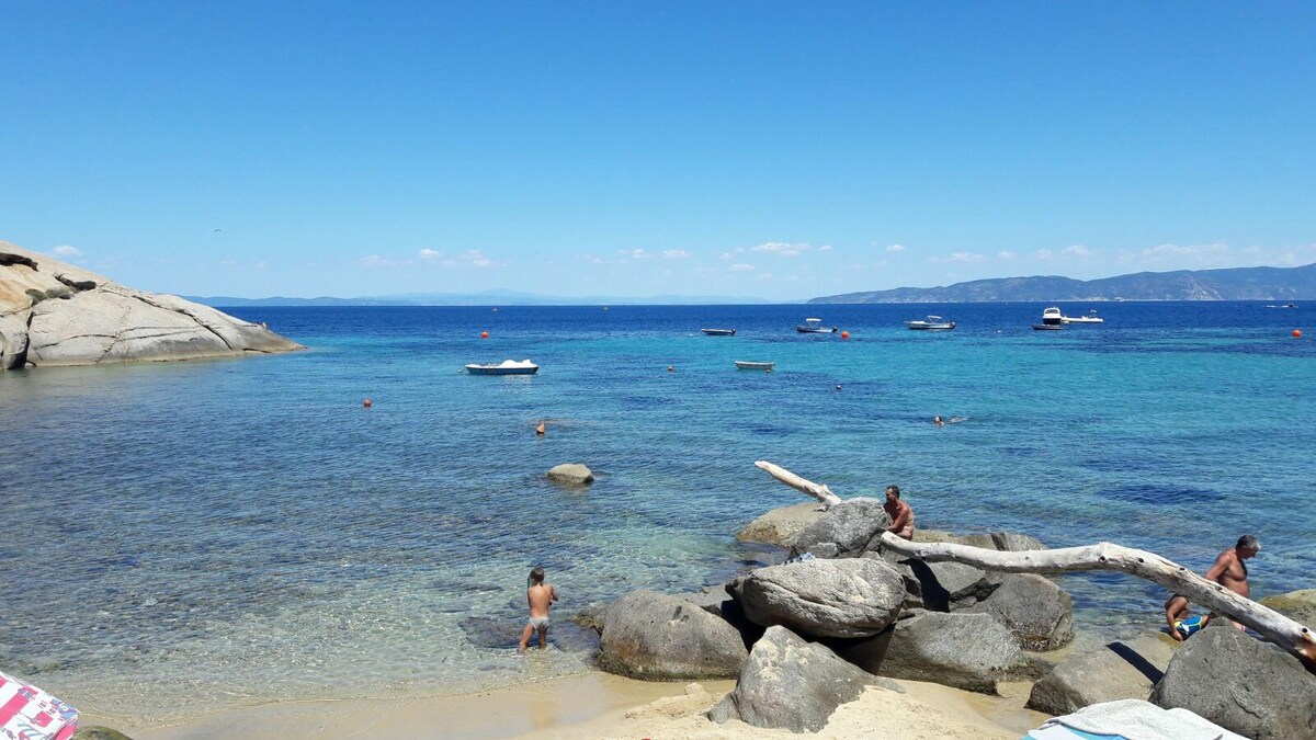 A sunny beach scene features clear blue waters with several boats floating in the distance. Rocky formations line the shore, while people can be seen enjoying the water and relaxing on the sand. The sky is bright and mostly clear, enhancing the coastal view.