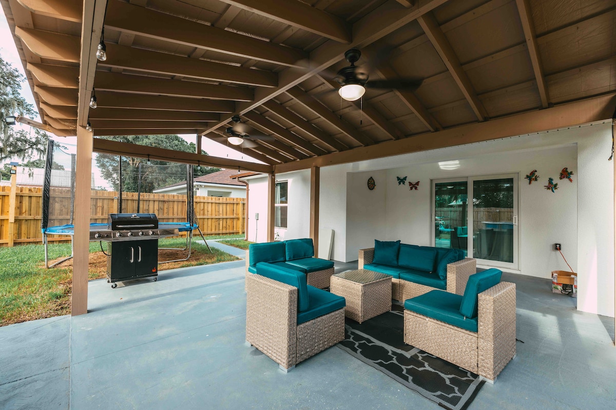 An outdoor patio area is presented with a comfortable seating arrangement, featuring a sectional and two chairs with teal cushions. A barbecue grill is positioned nearby. The space is sheltered by a roof, and the backyard is partially visible, showcasing a trampoline and a fenced area.