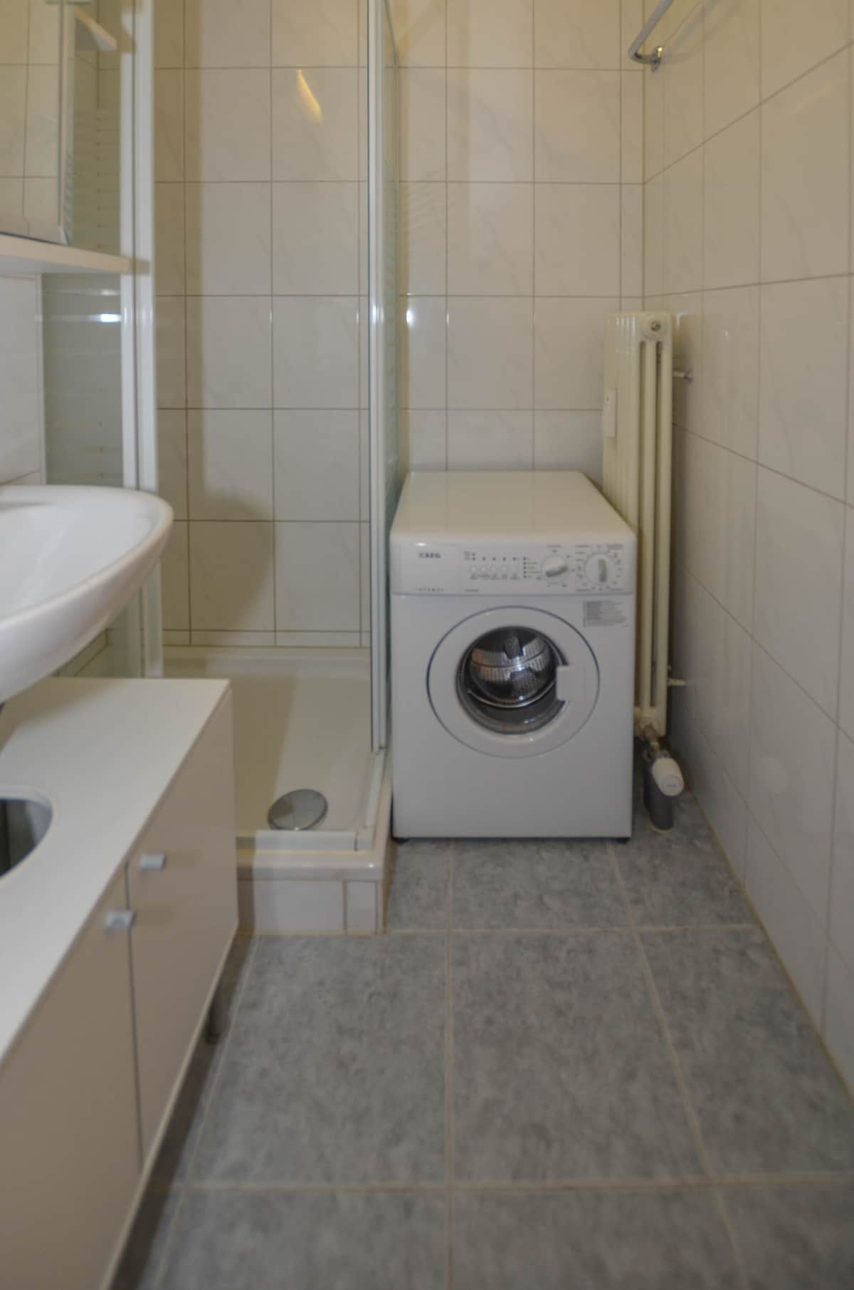 The bathroom features a glass shower enclosure and a washing machine positioned beside a white sink. Light-colored tiles cover the walls and floor, creating a clean and functional space. Shelving is provided for storage, and a towel radiator is mounted on the wall.