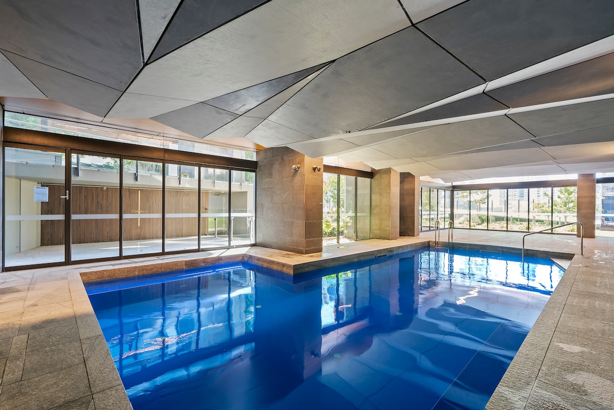An indoor swimming pool features a vibrant blue surface and is surrounded by large glass windows that bring in natural light. Sleek stone walls and an elegantly patterned ceiling contribute to a modern aesthetic. A pathway leads to the lush outdoor area visible through the windows.