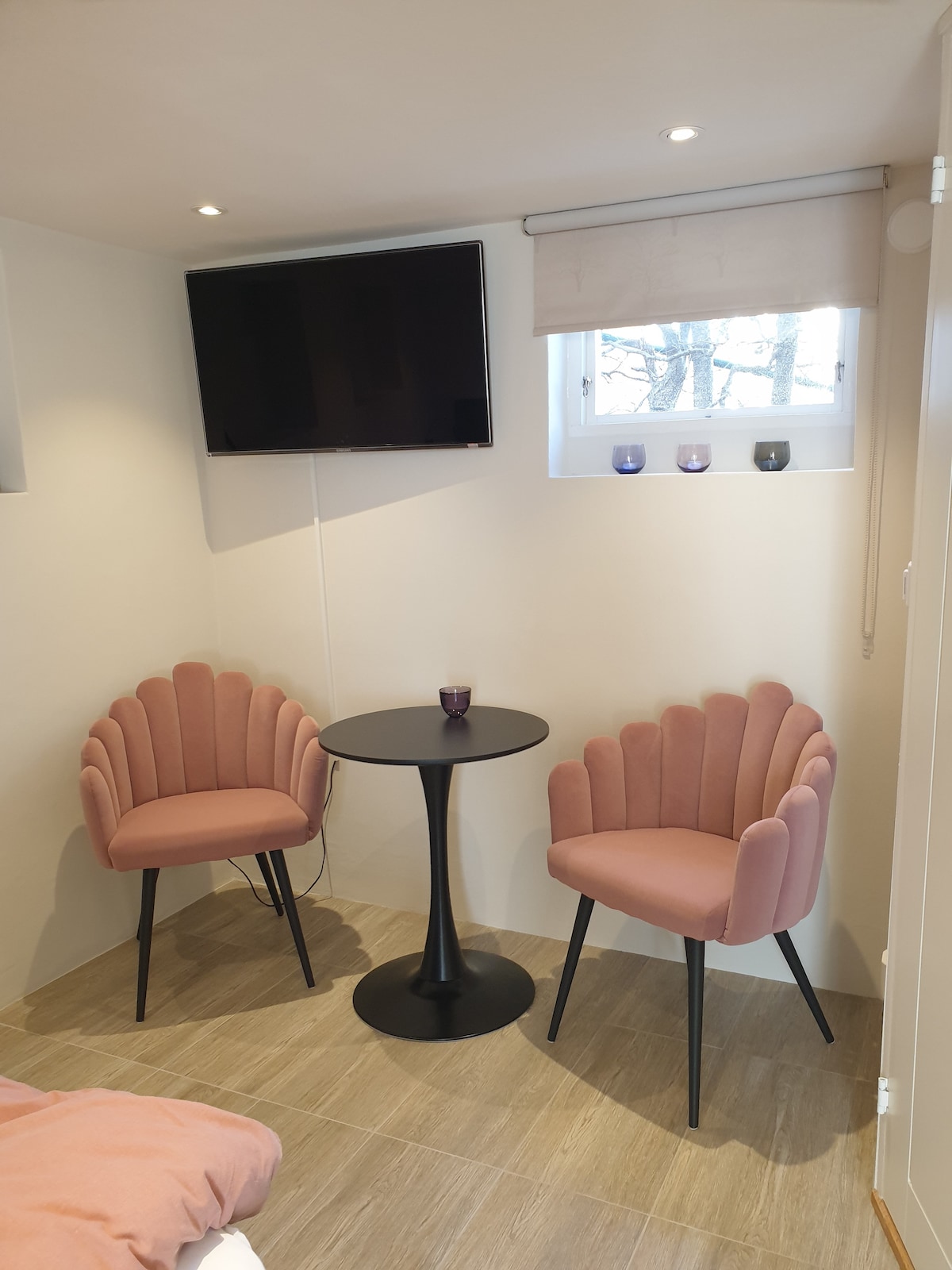 A cozy corner of the accommodation features two pink upholstered chairs styled with scalloped edges, positioned around a sleek black table. Above the table, a flat-screen TV is mounted on the white wall, with a window providing soft natural light. Three small candles are displayed on the windowsill.