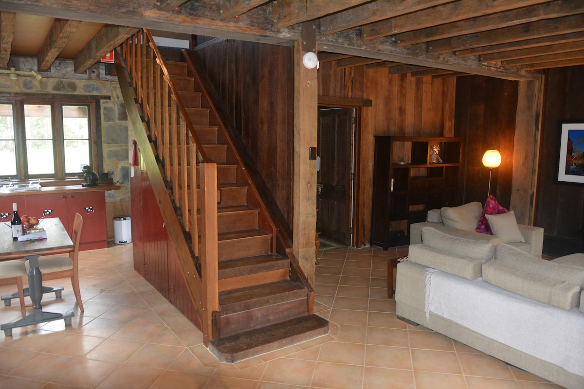 An open-plan living area is highlighted by exposed wooden beams and a warm color palette. A staircase, crafted from natural wood, ascends to the upper level. A cozy sofa invites relaxation, while a dining table and kitchen area are visible in the background.