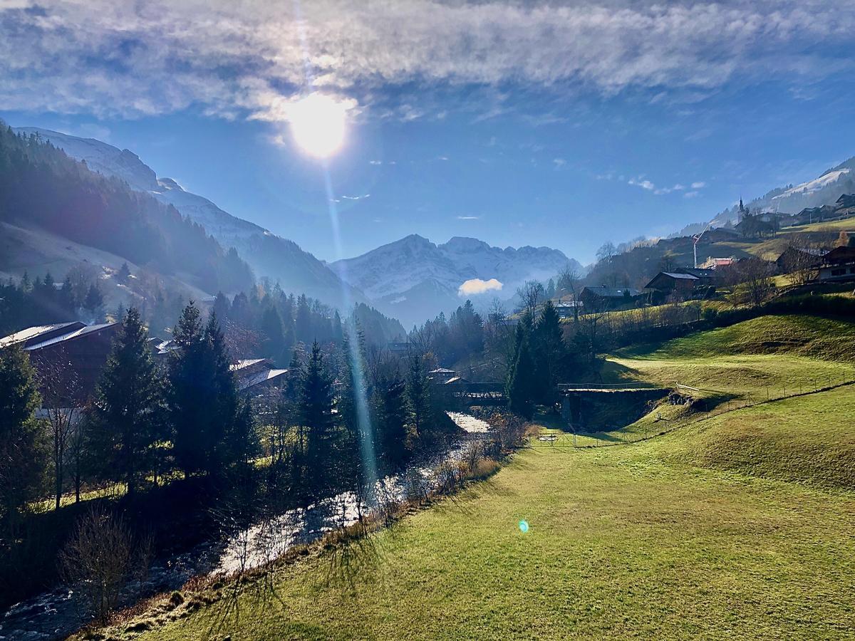 A panoramic view features rolling green fields and distant mountain ranges, bathed in natural sunlight. Trees are scattered throughout the landscape, with a serene stream meandering through. The scene captures the tranquility of the surrounding nature, enhancing the connection to the outdoors.