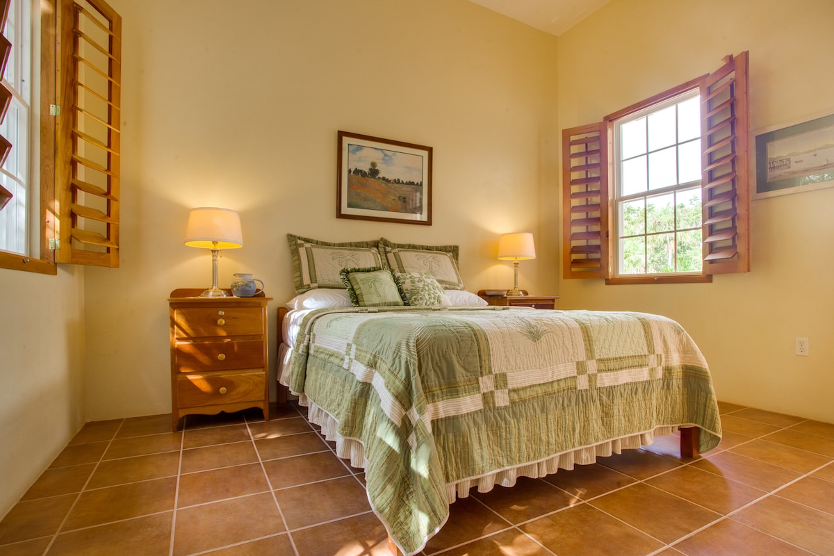 A well-lit bedroom features a spacious bed adorned with a quilted green coverlet. Wooden nightstands with lamps are positioned on either side. Windows with wooden shutters allow natural light to illuminate the terracotta tile floor and warm-toned walls.