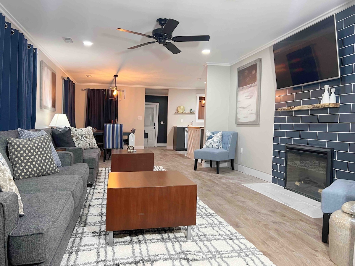 A contemporary living area is presented, featuring a comfortable gray sofa adorned with decorative pillows. A wooden coffee table is placed on a patterned area rug. A fireplace and wall-mounted television are visible, with warm lighting offered by ceiling fixtures and table lamps.