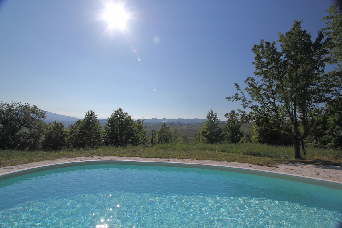 A private swimming pool reflects the sunlight, surrounded by a lush green landscape. Rolling hills and trees are visible in the distance, creating a serene and relaxing atmosphere. The clear blue water invites relaxation under the warm sun.