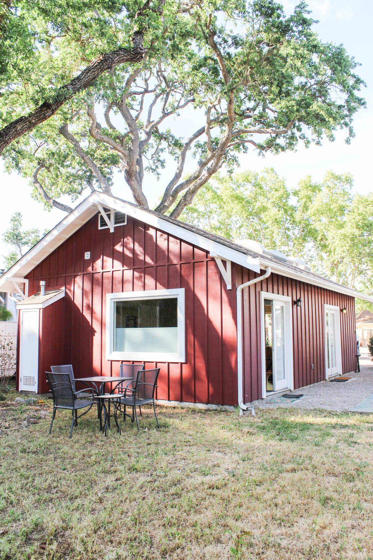 A red barn-style unit is set in a grassy area, framed by a large tree. A small outdoor table with four chairs is positioned in front, providing a space for relaxation. Large windows allow natural light to enter, while the entrance features white double doors.