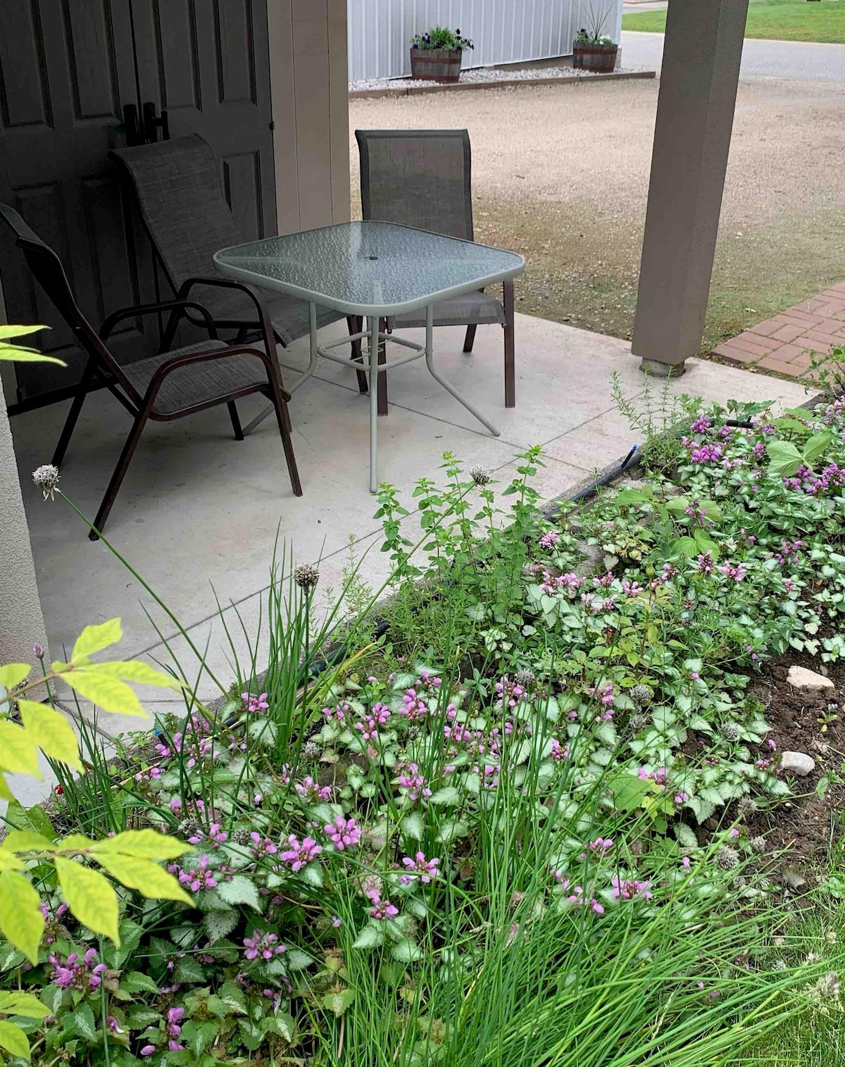An outdoor seating area is depicted, featuring a glass-top table accompanied by two chairs. Lush greenery and blooming flowers surround the space, enhancing the natural setting. The entrance is sheltered by a roof, providing a comfortable area for relaxation.