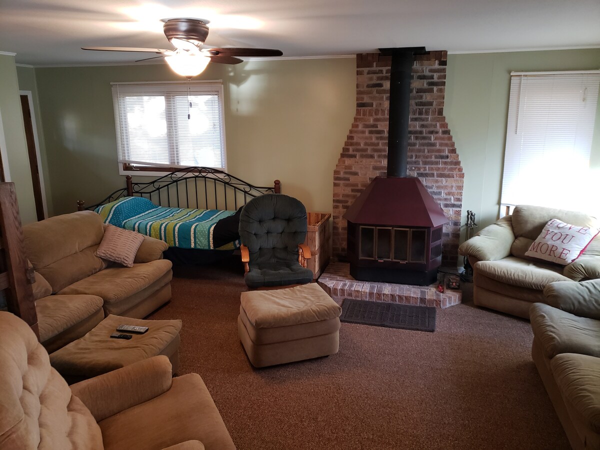 A spacious living area is arranged with multiple comfortable seating options, including two sofas, a large armchair, and a cozy ottoman. A stone fireplace with a distinct brick surround serves as the focal point, complemented by a bed and colorful blanket positioned nearby.