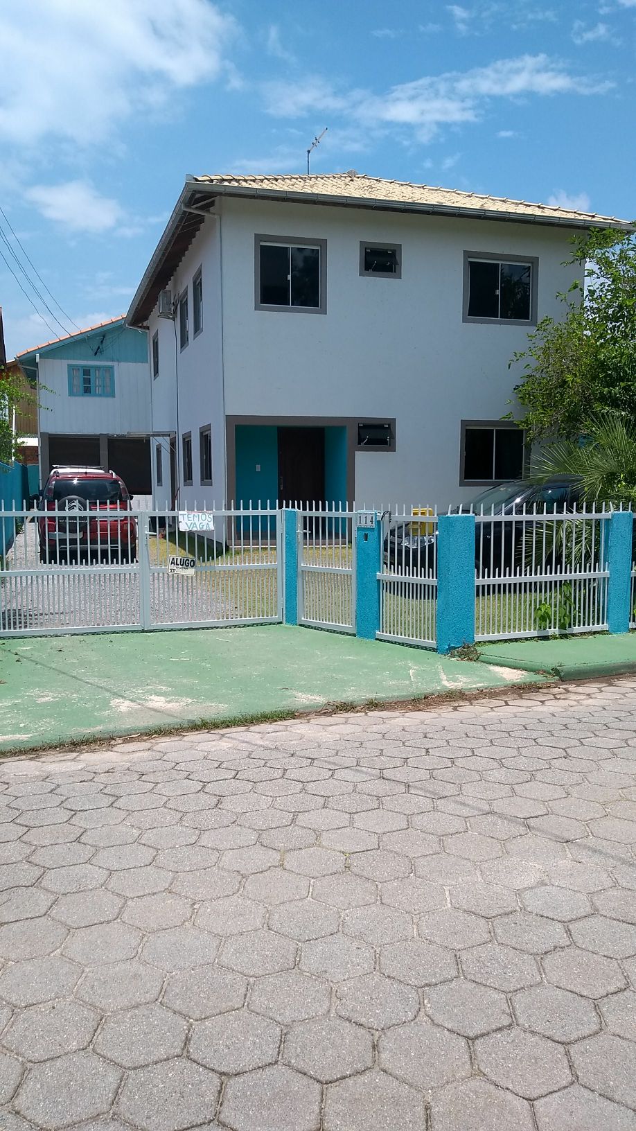 A two-story building is surrounded by a fenced yard with a gate. The structure features large windows and a light-colored façade. A paved area accommodates vehicles, with several parked cars visible. Lush greenery adds to the inviting exterior on a sunny day.
