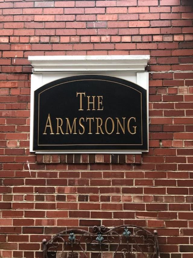 A black sign with gold lettering displays the name 'The Armstrong.' The sign is mounted on a textured red brick wall, which adds a classic architectural element to the setting.